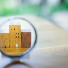 A magnifying glass focuses on three wooden house-shaped blocks arranged on a table, symbolizing home inspections, real estate, or property evaluation. The background is softly blurred with natural light and greenery.
