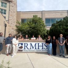 Group photo of 10 RMHS staff members standing outside the Rocky Mountain Human Services building, with the RMHS sign displayed in front of them.