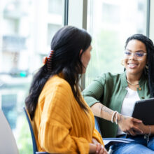 Two women sit together in an office setting, engaged in a conversation. One woman holds a tablet while smiling and listening to the other woman.