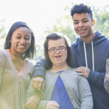 Five diverse young adults stand together outdoors, smiling and laughing, with one young woman with Down syndrome standing in the center.